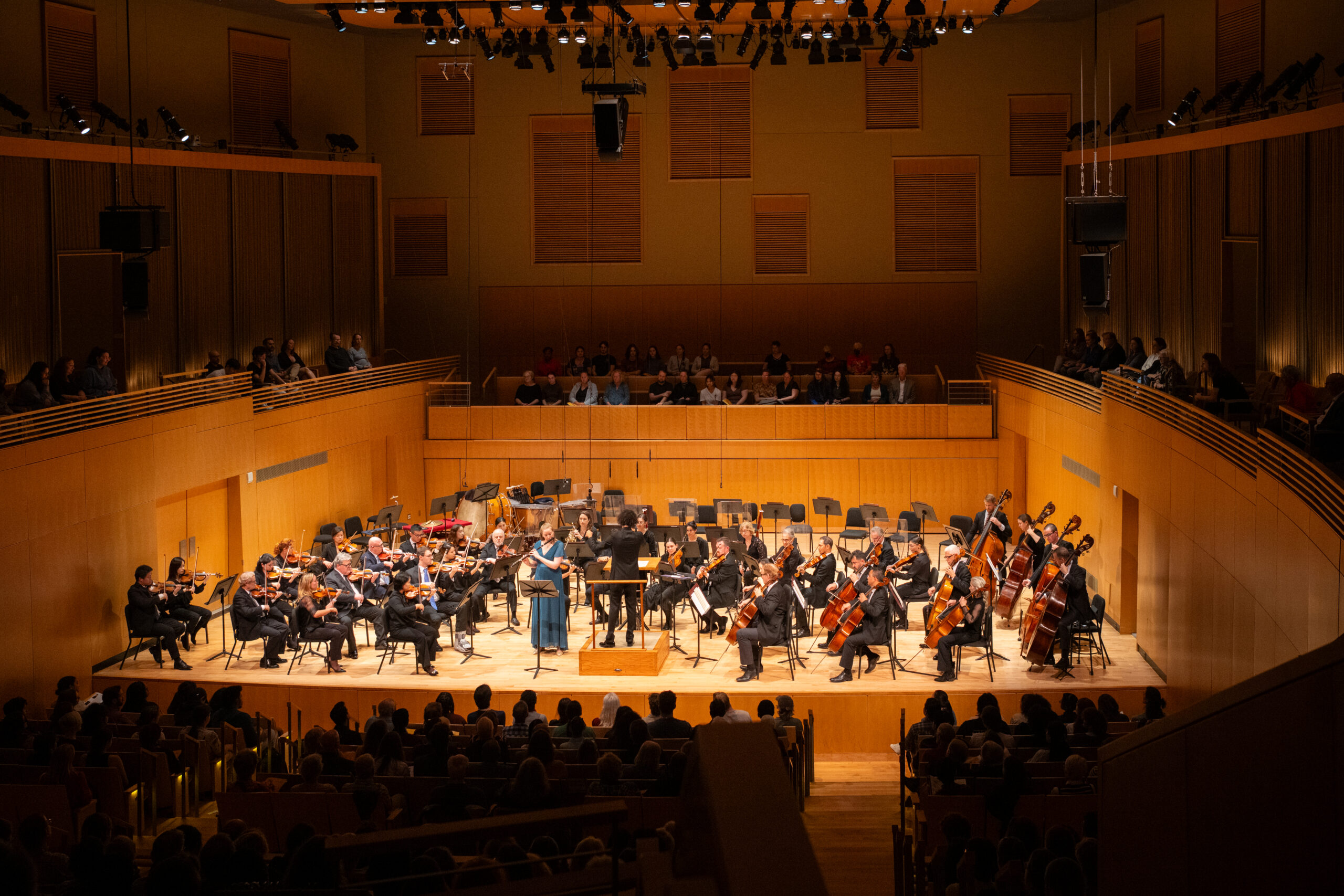 A far away view of the Baltimore Symphony Orchestra performing onstage with their conductor. The audience watches from all angles.