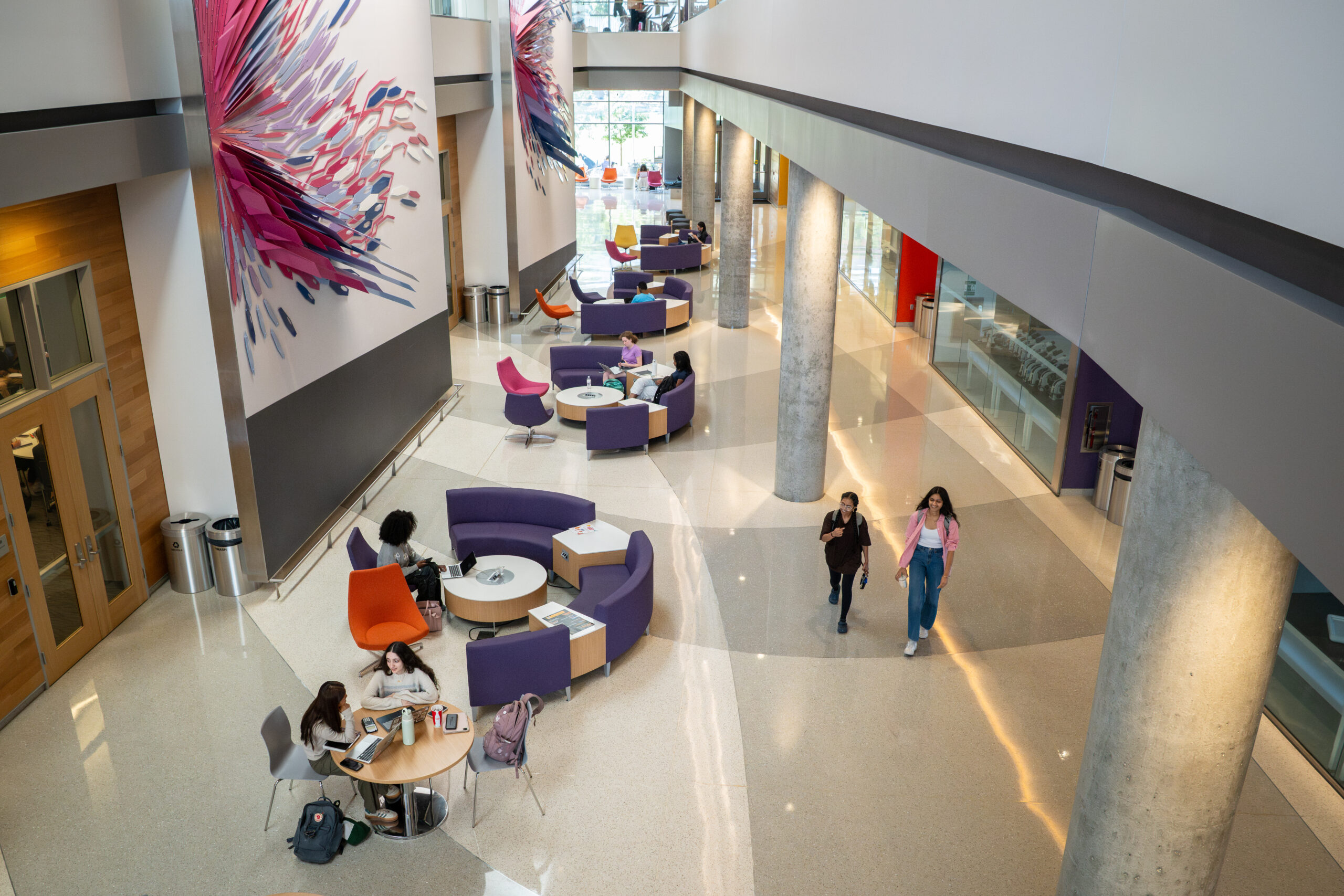 Students sit at tables and walk through hallway. Columns punctuate the hallway and colorful art is displayed on the walls.