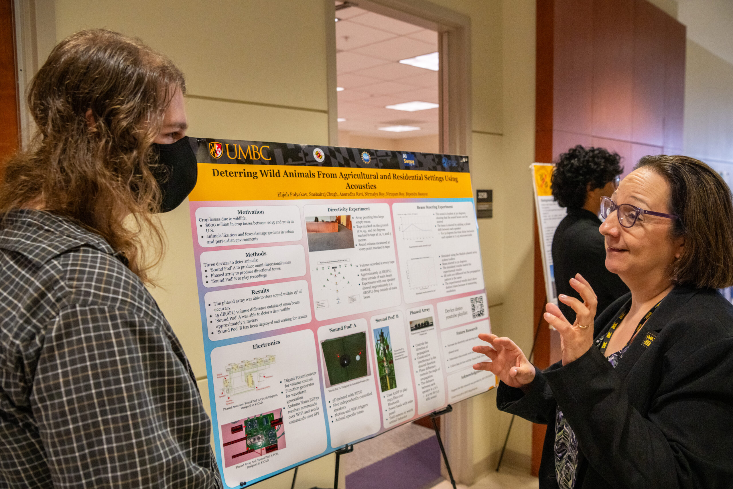 Student in mask talks with woman in jacket and glasses in front of scientific research poster.
