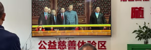 A group of men and women look at a big screen with men talking on it.