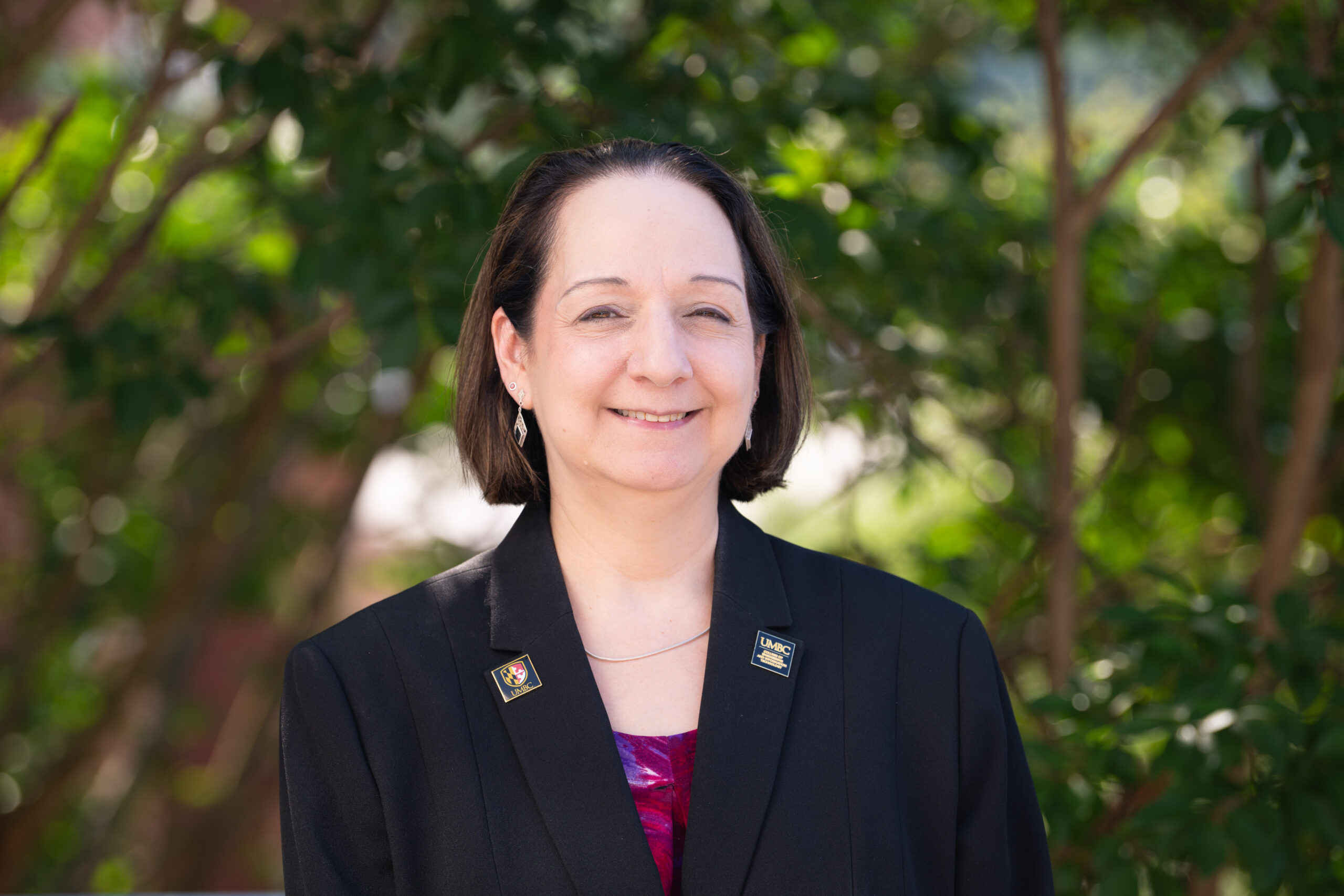 Jeanne van Briesen in black jacket with UMBC pins on the lapels smiles at the camera.