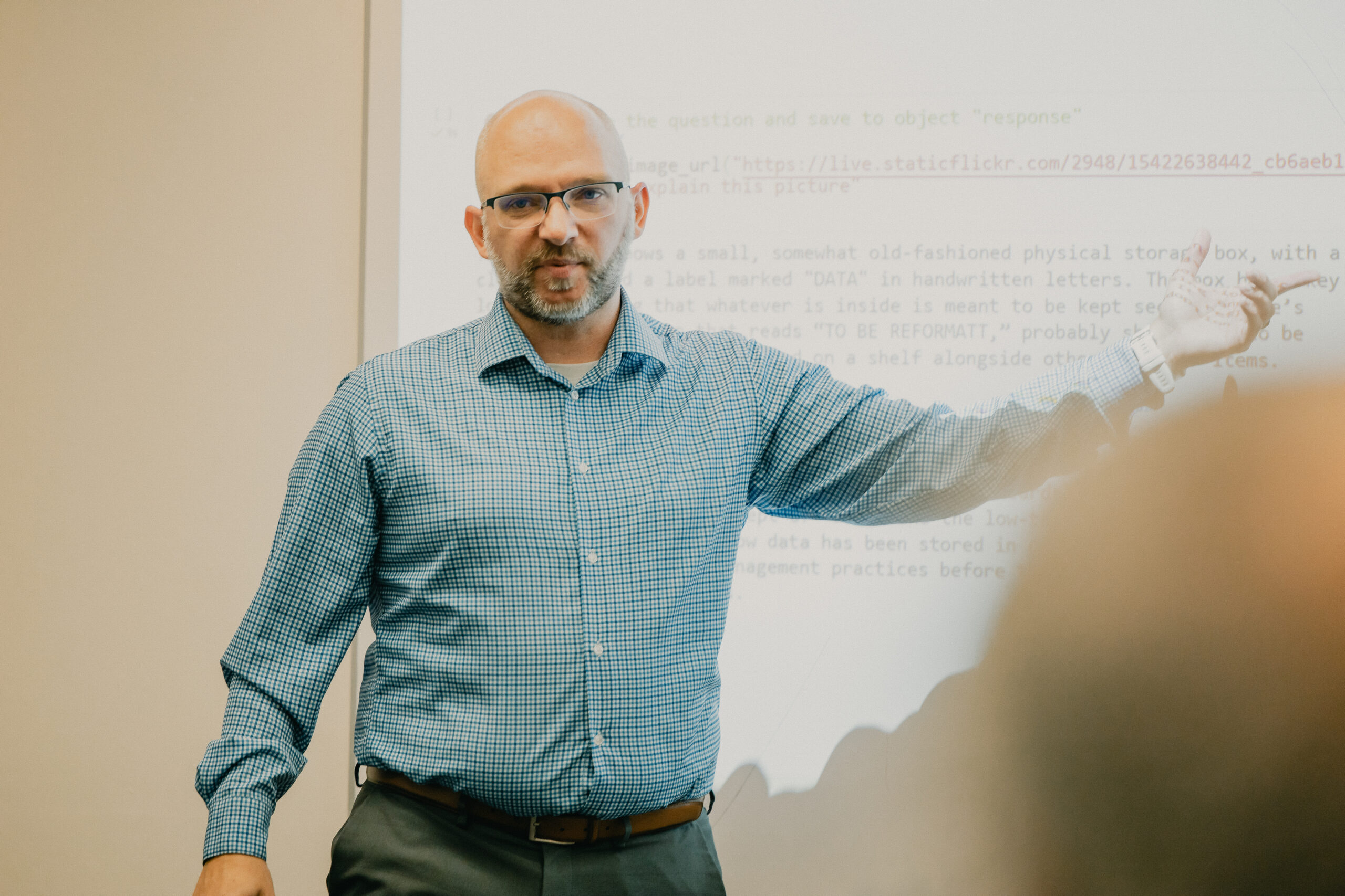 Eric Stokan, college professor wearing a checkered blue and white dress shirt points to a project screen with computing code language