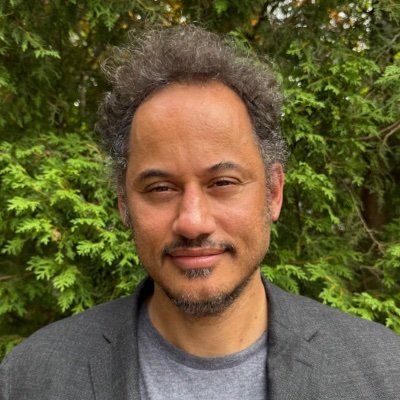 A man with curly hair wearing a grey shirt, looking directly at the camera with a neutral expression.
