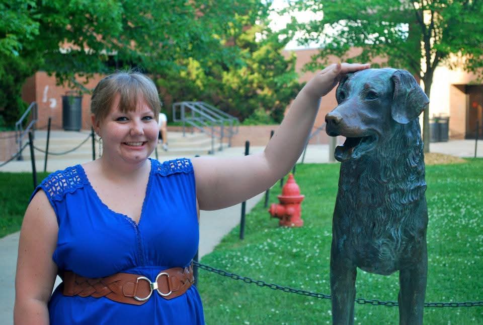 A woman in a blue dress stands next to a dog statue, smiling and patting its head.