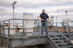 man stands on metal paltform on the roof of a building; several large scientific instruments sit behind him on the platform