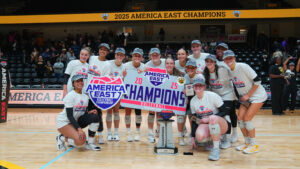 UMBC's volleyball team huddled holding up #1 fingers around the America East trophy with America East champion hats on and holding the America East sign
