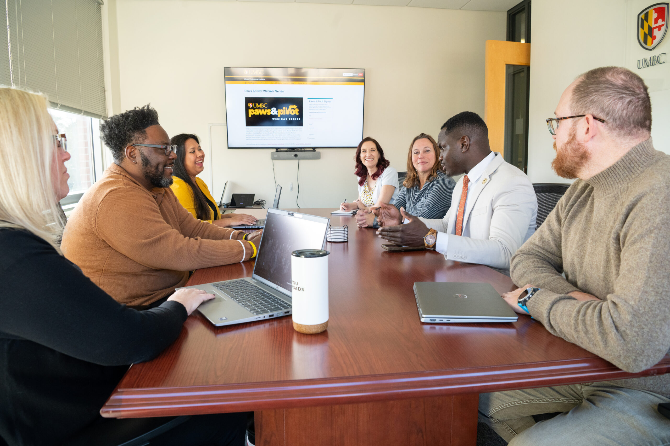 A group of people gather around a boardroom table with a TV screen in the background with the words Paws & Pivot