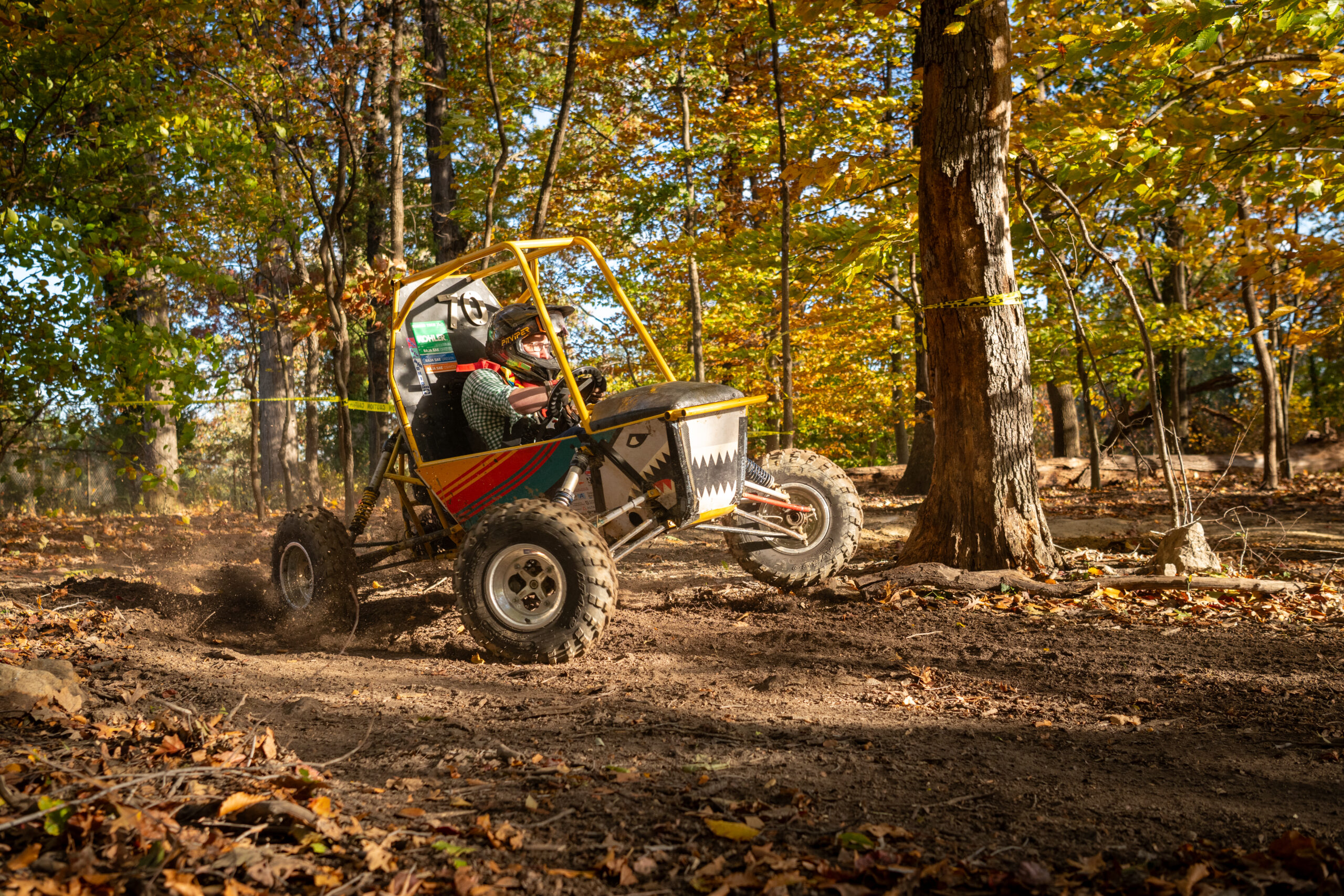 A driver in a one-seat off-road vehicle kicks up dirt as they round a turn on a dirt path through the trees.