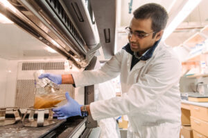 grad student in lab coat, gloves, and safety glasses handles a large flask under a fume hood containing a tan liquid