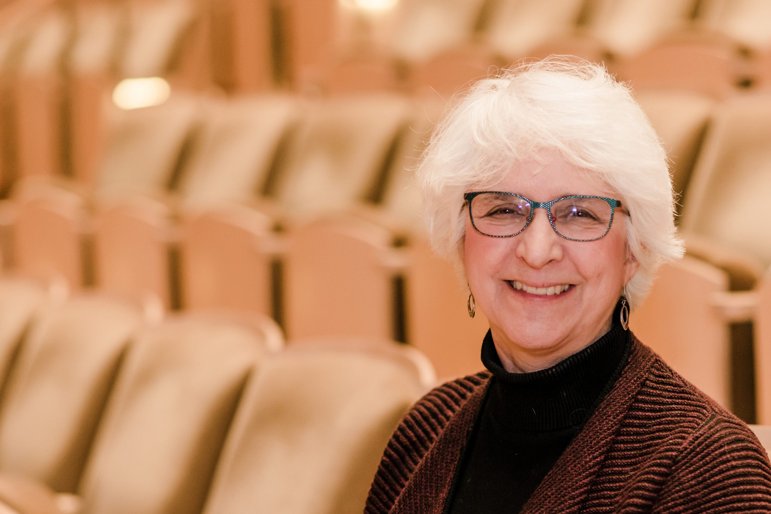 Linda Dusman, who has white hair, wears glasses, and wears a brown and black outfit.