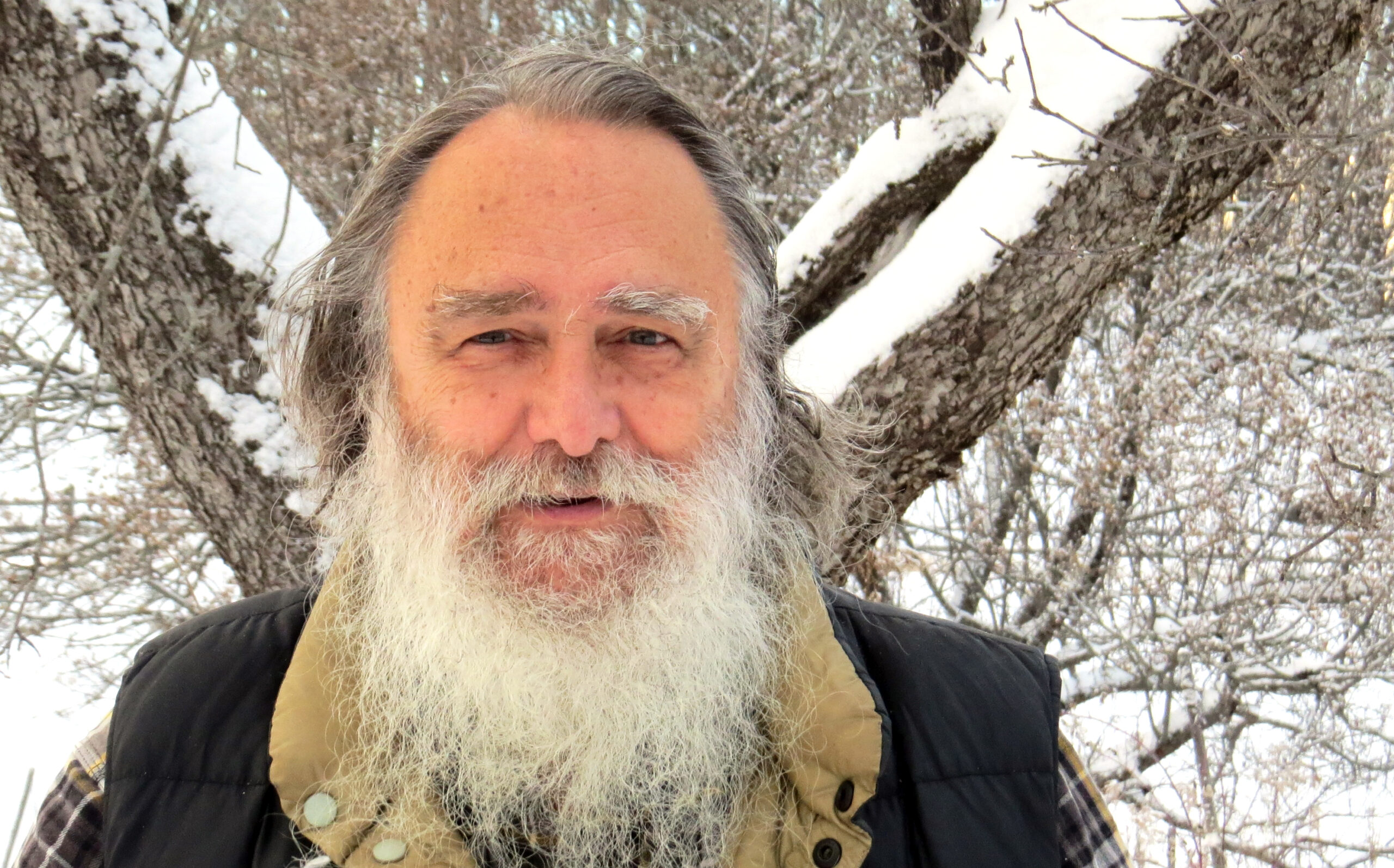 Against a wintery background, a white man with dark hair and a white beard looks at the camera.