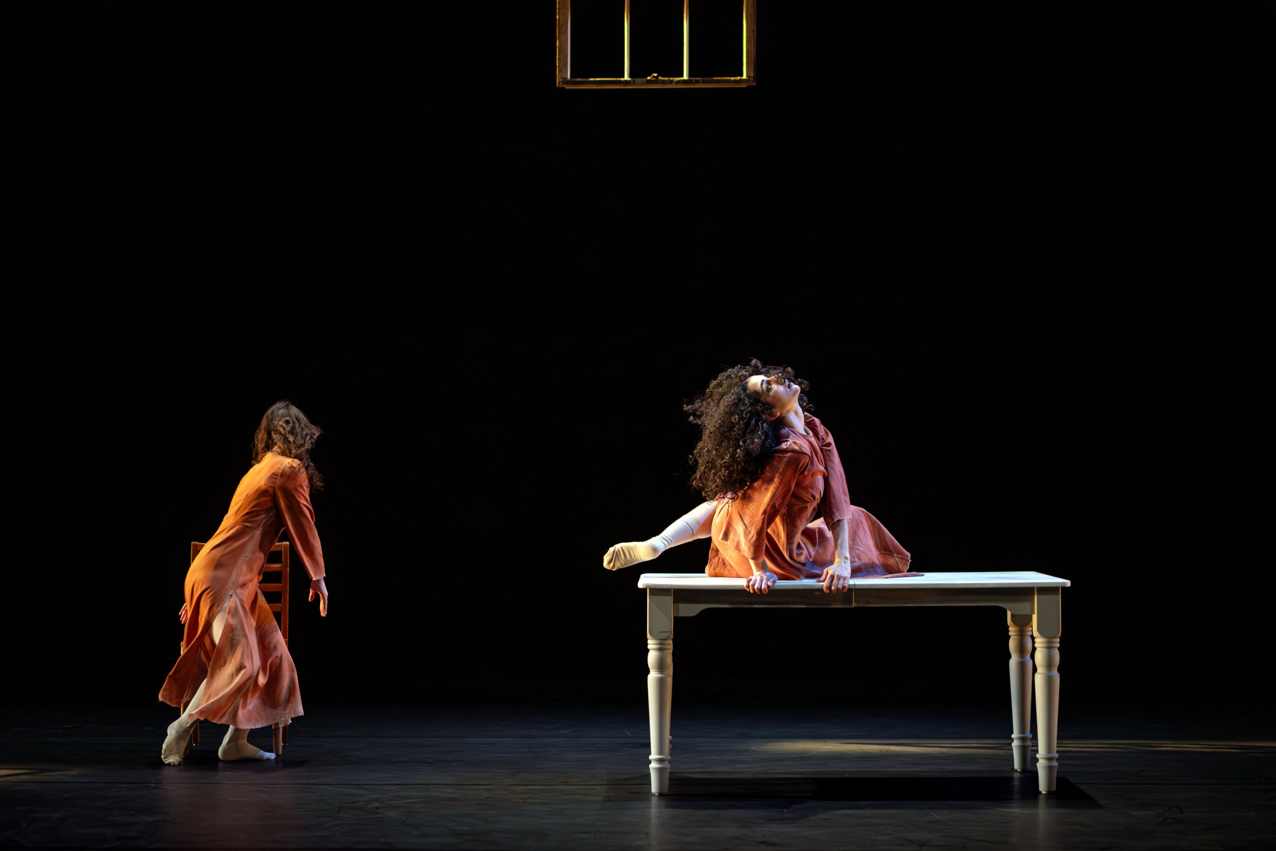 Against a black background, we see two dancers on stage . On the left a dancer is turning toward a chair, and on the right a dancer is on top a table, looking up.