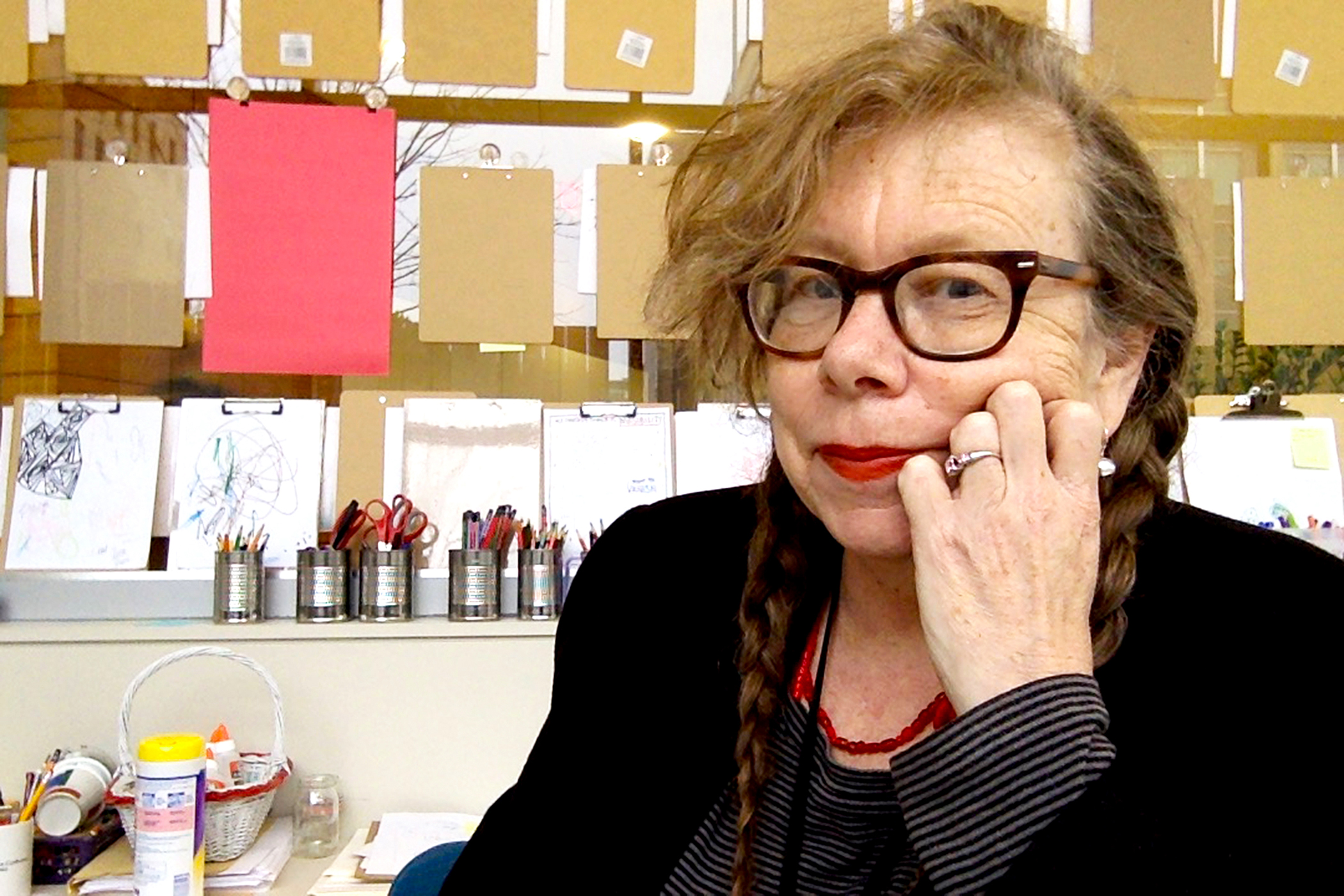 Against a background of art sketches, a woman with strawberry hair and glasses, wearing a dark top, rests her chin in her hand and looks at the camera.