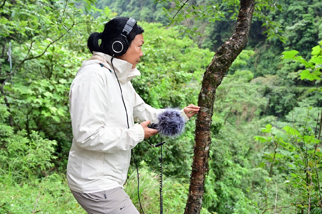 Against a lush forested background, a woman in profile who wears a white jacket and headphones, holding a microphone.