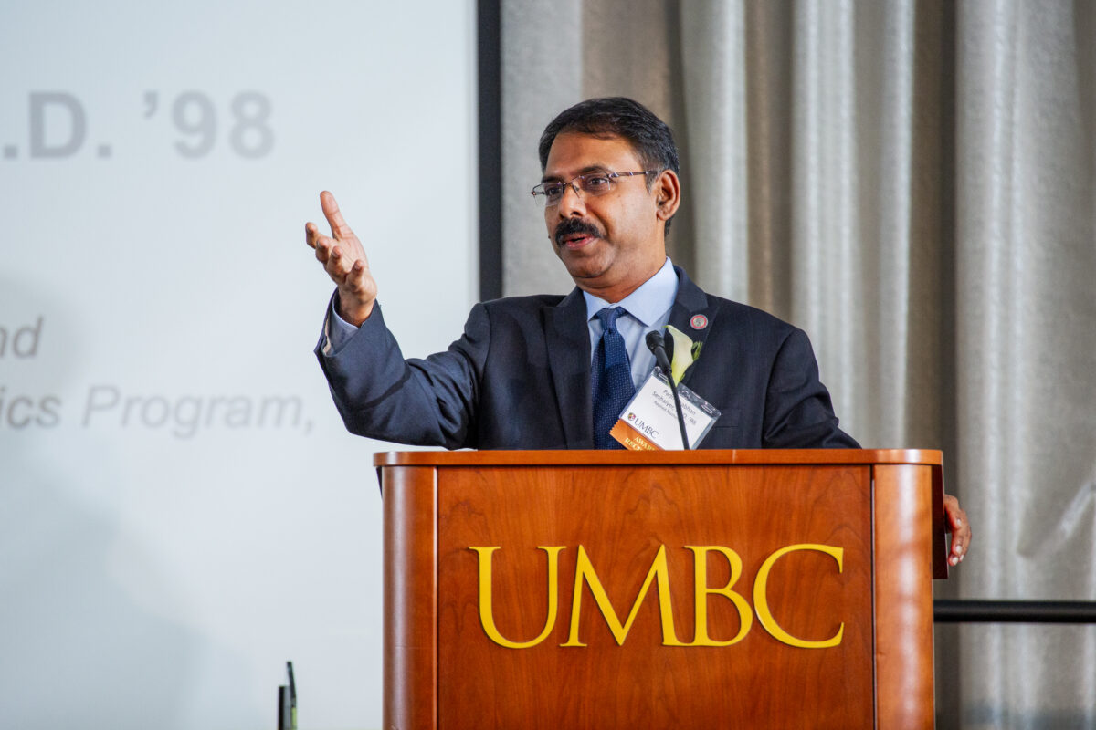 man stands at lectern that reads "UMBC" in gold letters, looking out and one arm extended