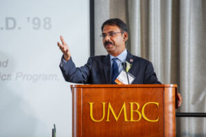 man stands at lectern that reads "UMBC" in gold letters, looking out and one arm extended