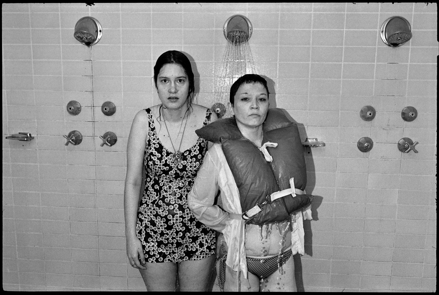In a black and white photograph, two clothed women stand side-by-side in a shower, the one on the right wearing a life vest.