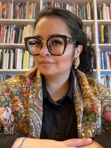 Against shelves of books, a woman with dark hair and glasses, wearing a colorful top, looks at the camera.