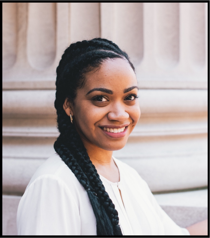A Black woman will long braided hair, wearing a white, top, smiles at the camera.