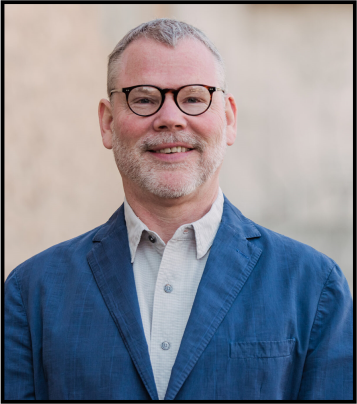 A white man with short gray hair and slight beard, wearing glasses and a blue jacket, smiles at the camera.