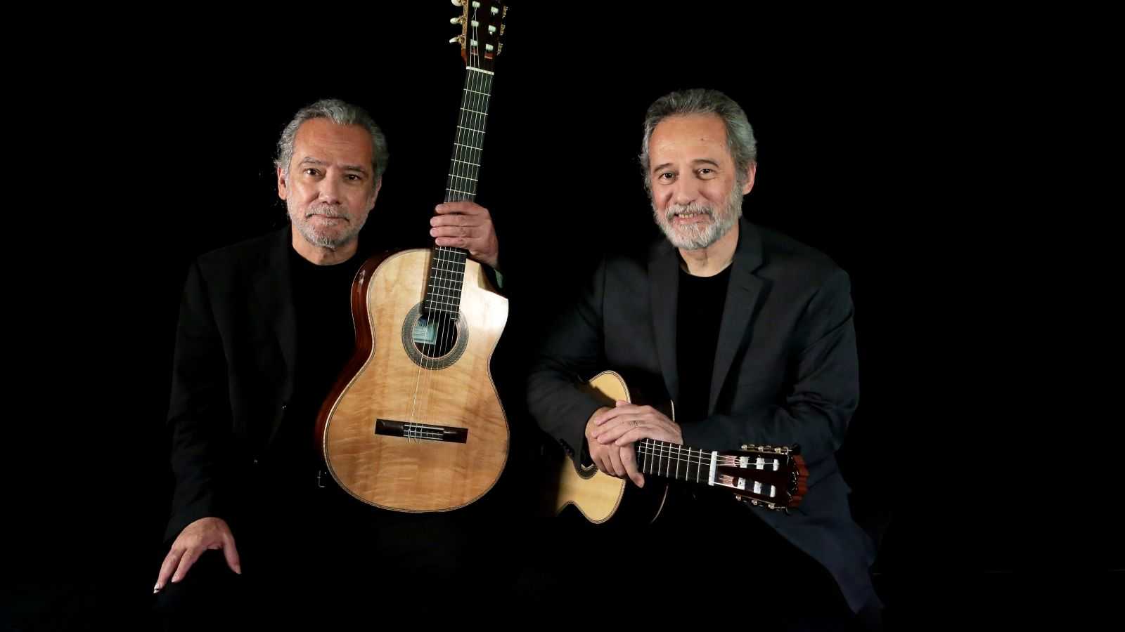 Against a black background, two men, both with gray hair and slight beards, hold classical guitars and smile at the camera.