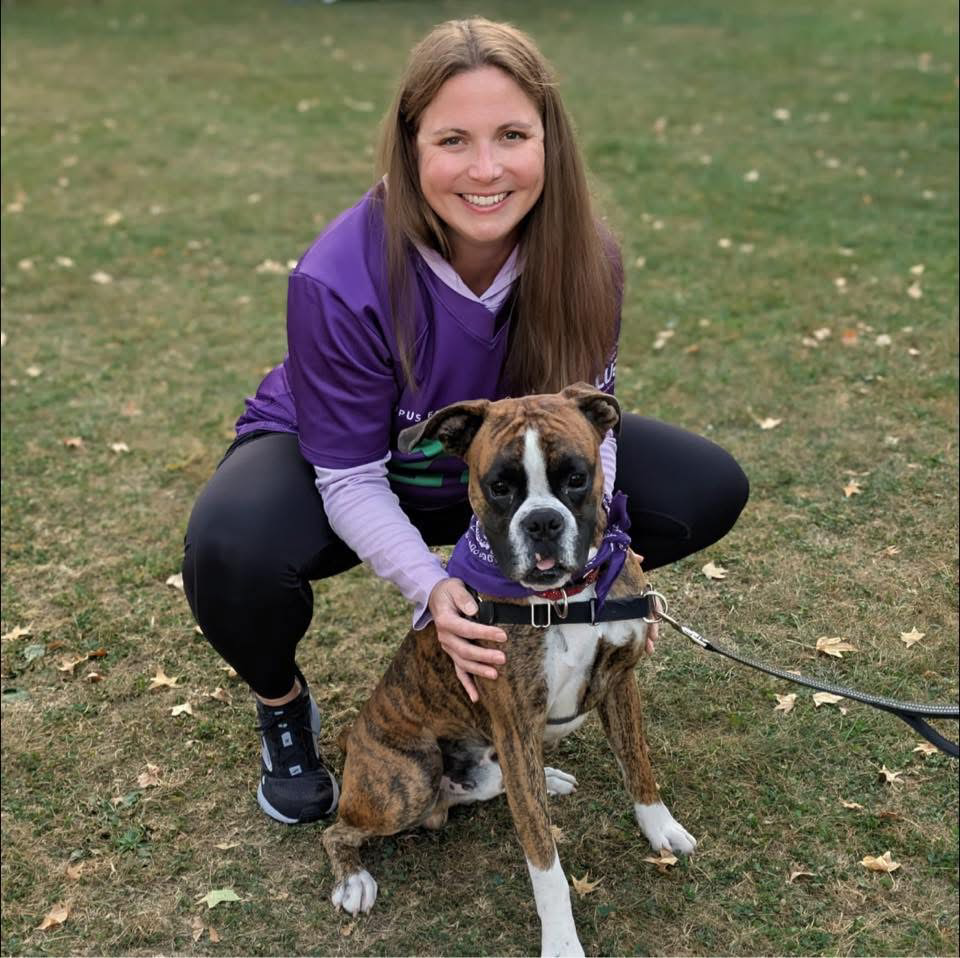 woman in purple t-shirt branded with the Lupus Foundation of America, which supports people with the chronic illness, squatting on a grassy field with her hands on her brown and white dog