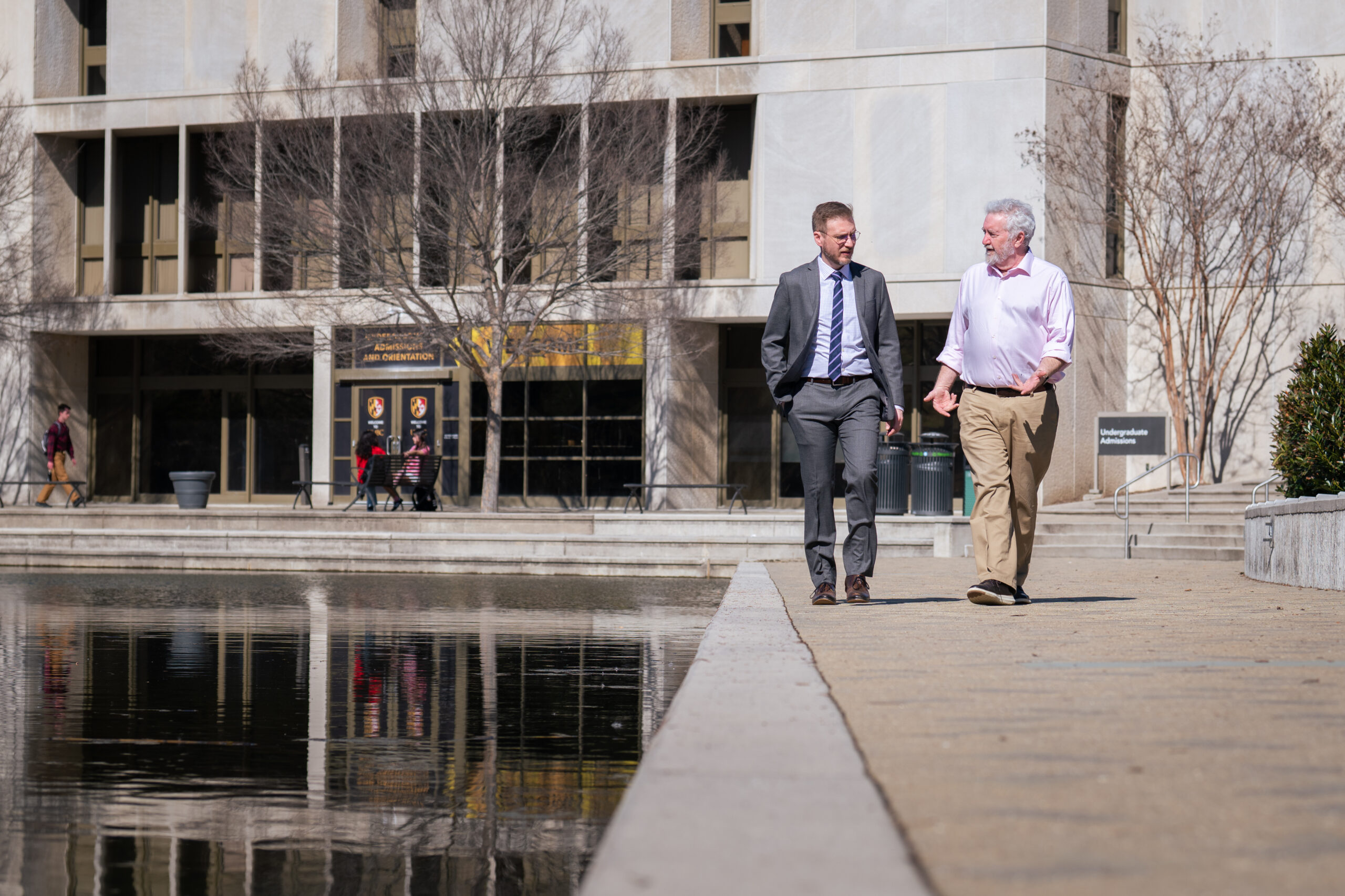 two men in conversation walk alongside the UMBC Library Pond, which serves to help prevent flooding on campus