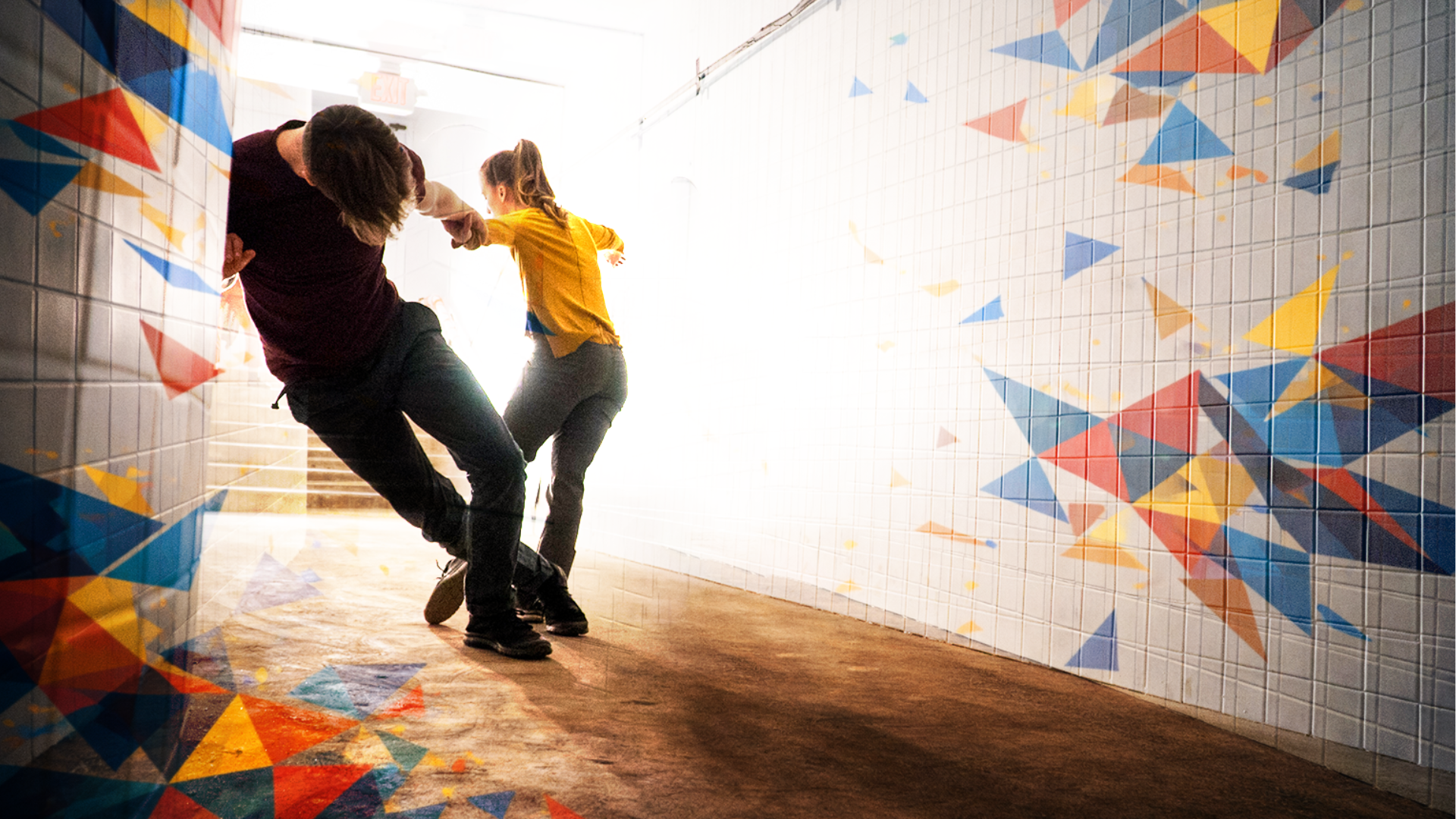 In a vividly lit corridor, a man in black dances with a woman in a yellow top.