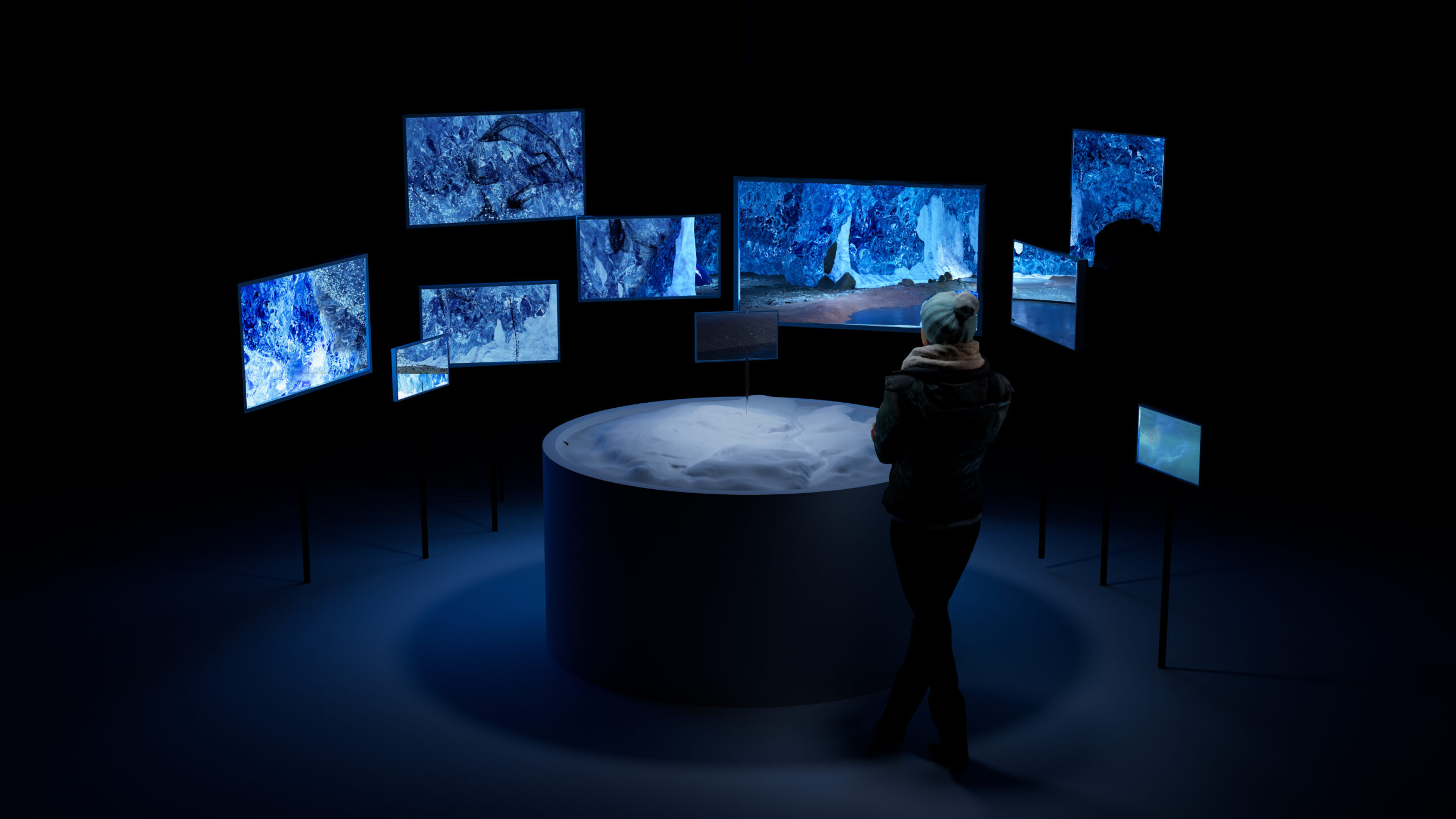 In a dark and dimly-lit space, a person stands near a cylindrical object on the ground, surrounded by flat screens that are omitting blue images.