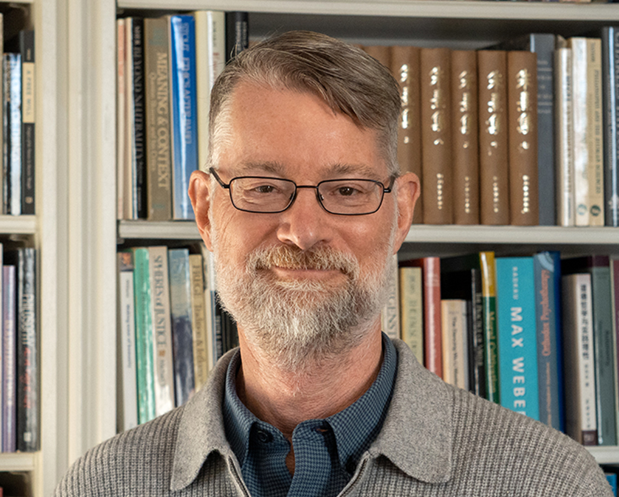 A white man with glasses and a slight beard, who poses in front of a bookshelf and smiles.