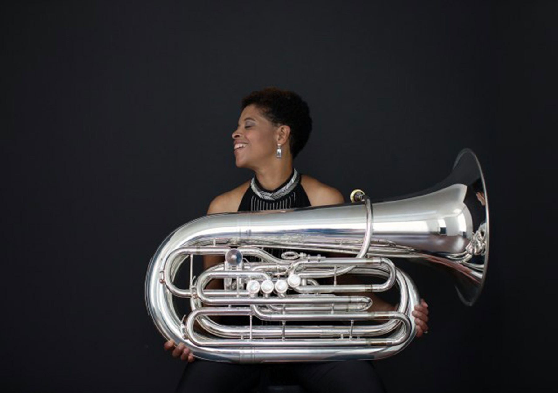 Against a dark background, a Black woman holds a tuba.