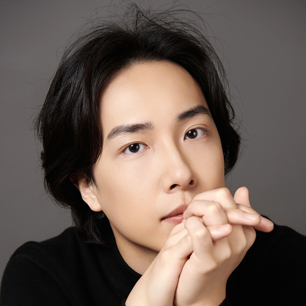 A headshot of a man with a medium skin tone and dark hair. He wears a black top, and clasps his hands at his chin, looking at the camera.