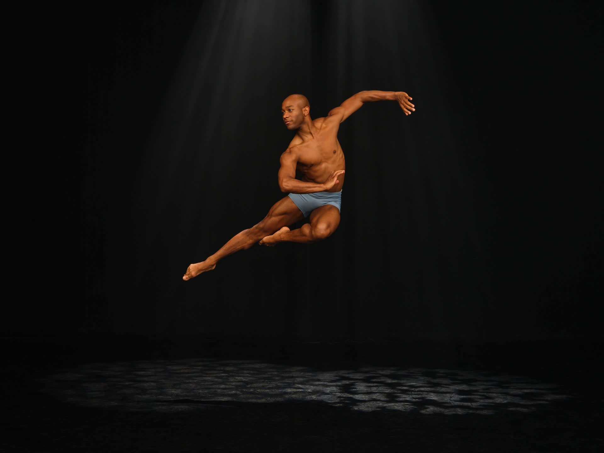 Xavier Mack jumping in dance pose in a spotlight on a dark stage.
