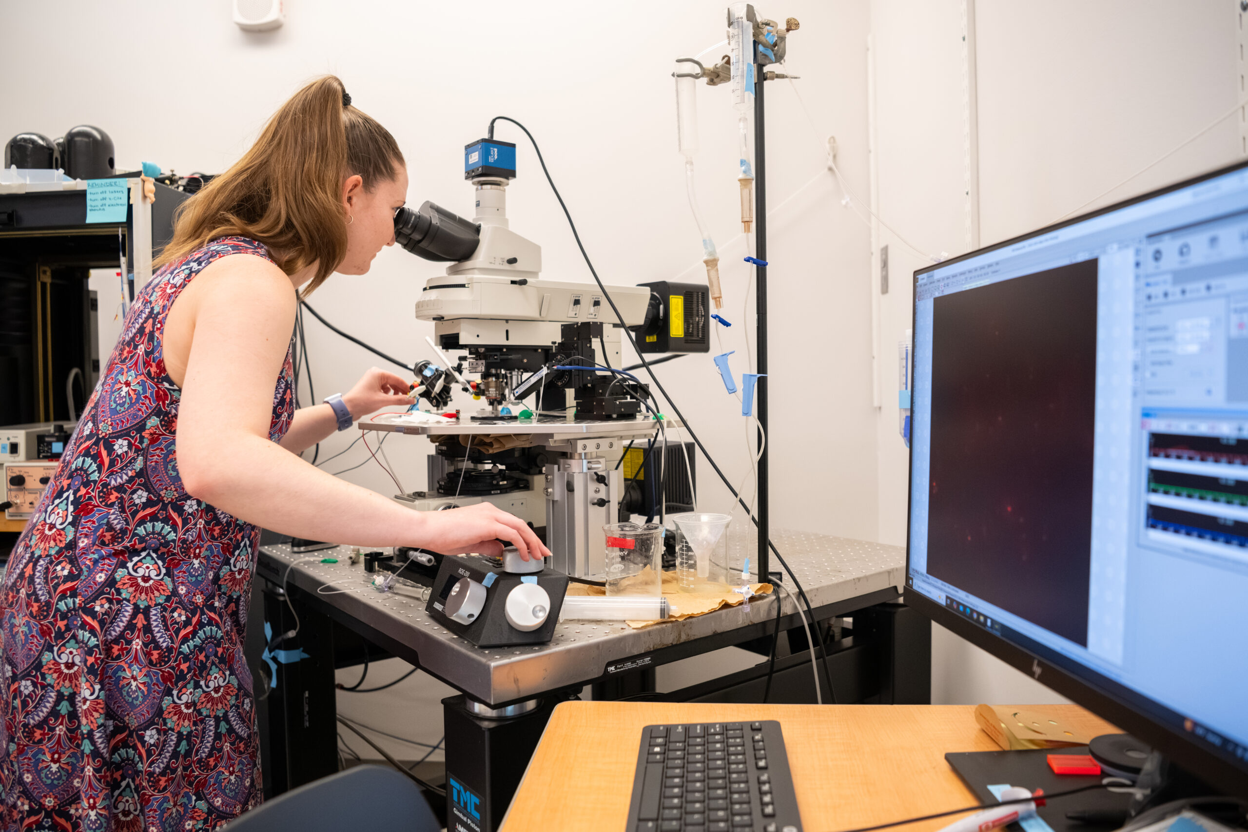 woman in sleeveless floral dress stands looking into microscope; computer monitor to the right shows data