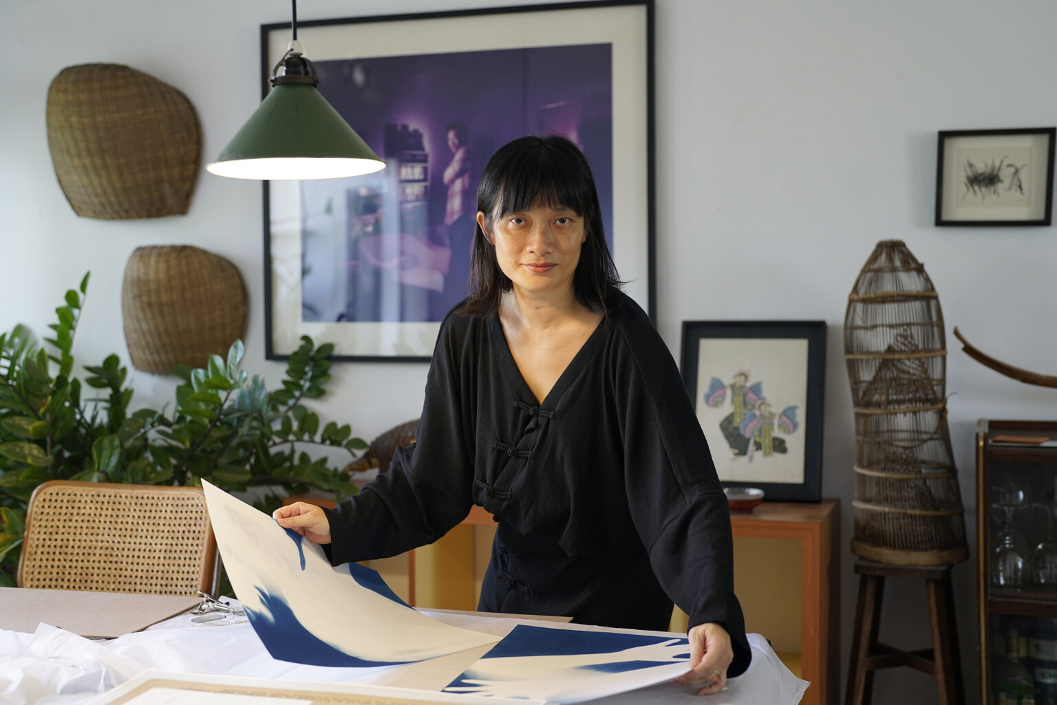 A woman with a medium skin tone, black hair, and black outfit, holds a piece of artwork as she looks at the camera.