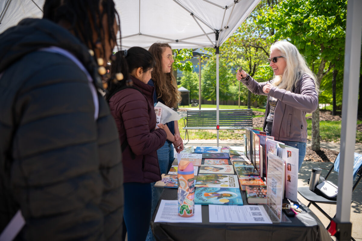 people converse under an EZ-up across a table covered with book