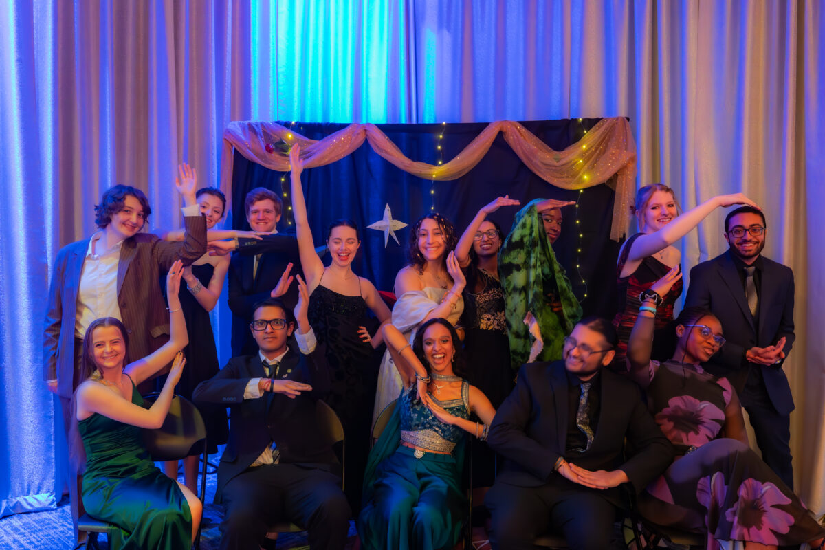 Several students in formalwear strike poses in front of a dark backdrop with ribbons