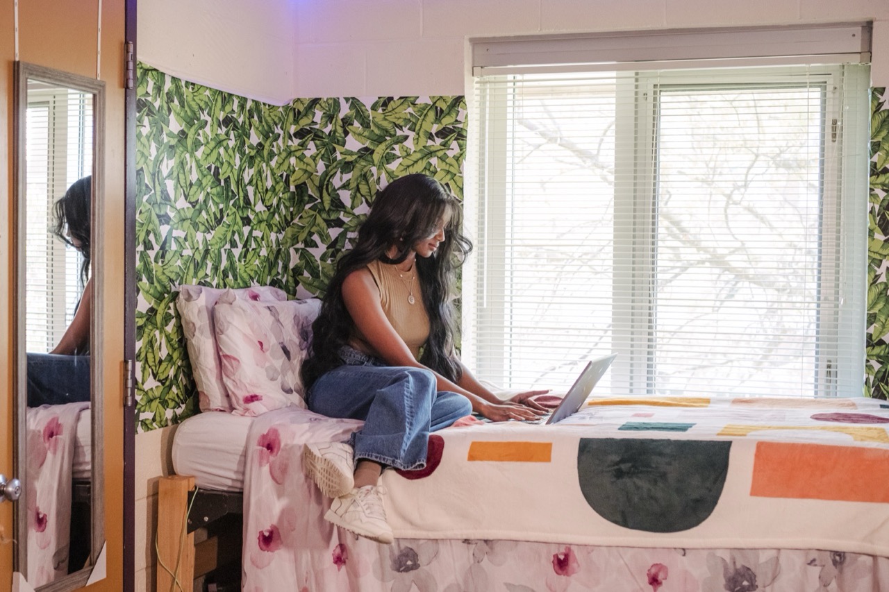 In a dorm room, a person sits on a bed using a laptop near a window.