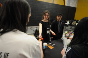 A girl, a member of the UMBC Honors College, stands with her arm outstretched behind a table showing art work.