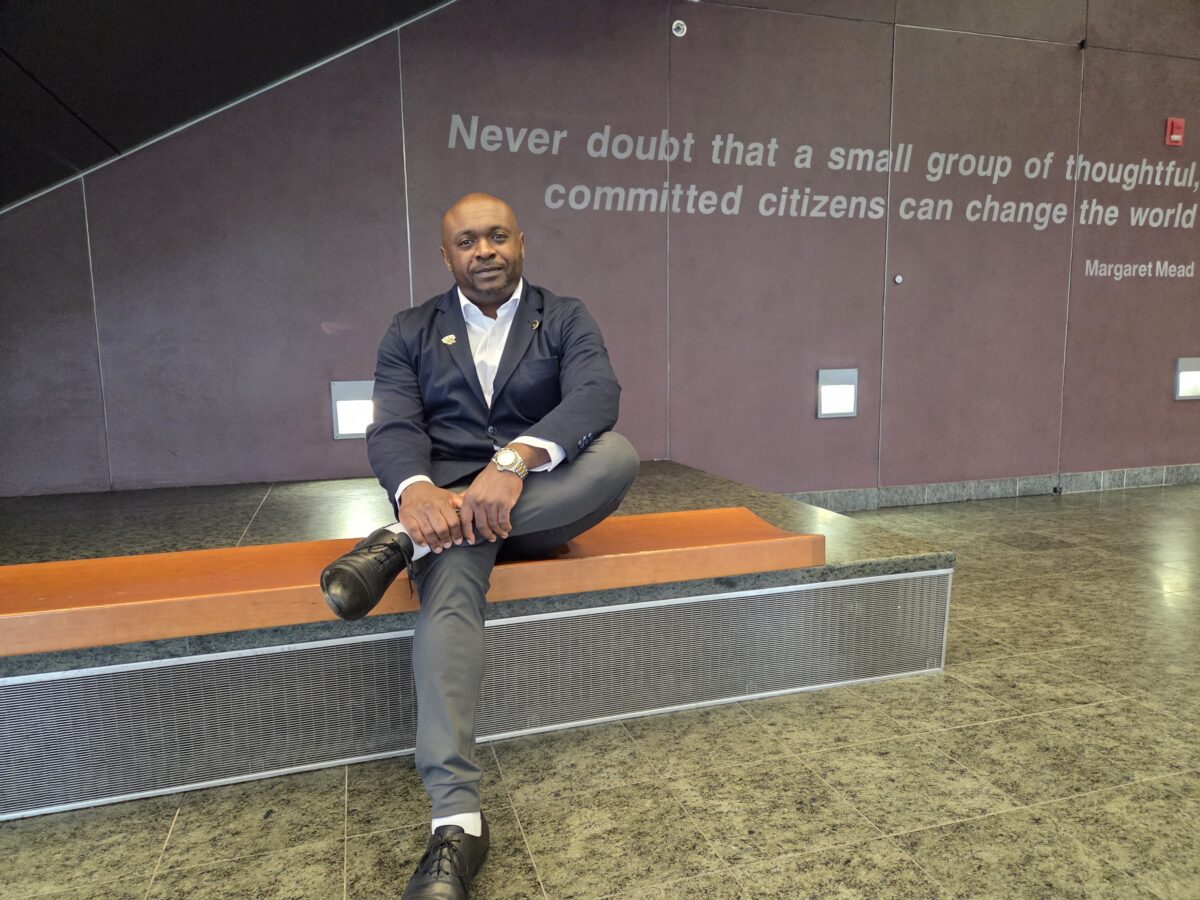 Demetrie Garner, a doctoral student, sits in a lobby of a building on a wooden bench inside in front of a wall with the quote "Never doubt that a small group of committed citizens can change the world, opioid abuse disorder
