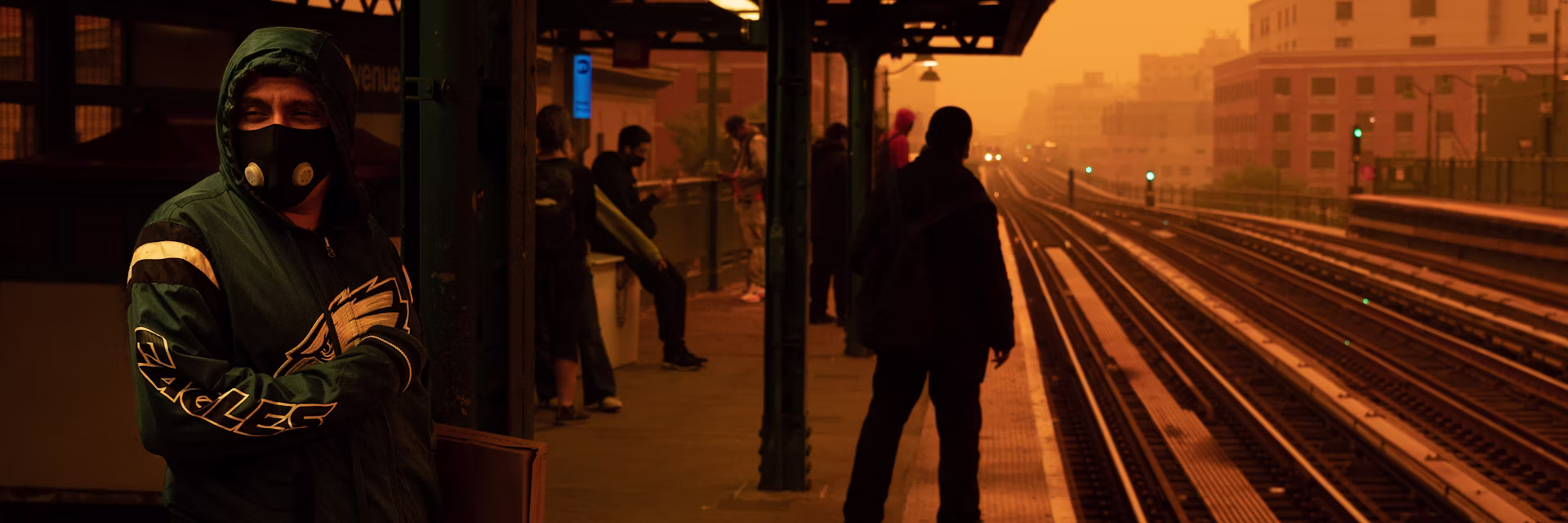 people waiting for a train wearing masks; the sky is yellow-orange