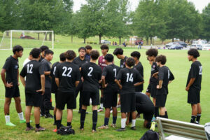 A soccer team wearing black uniforms with white numbers huddle around their coach, Balmory Moran, while standing on a soccer field