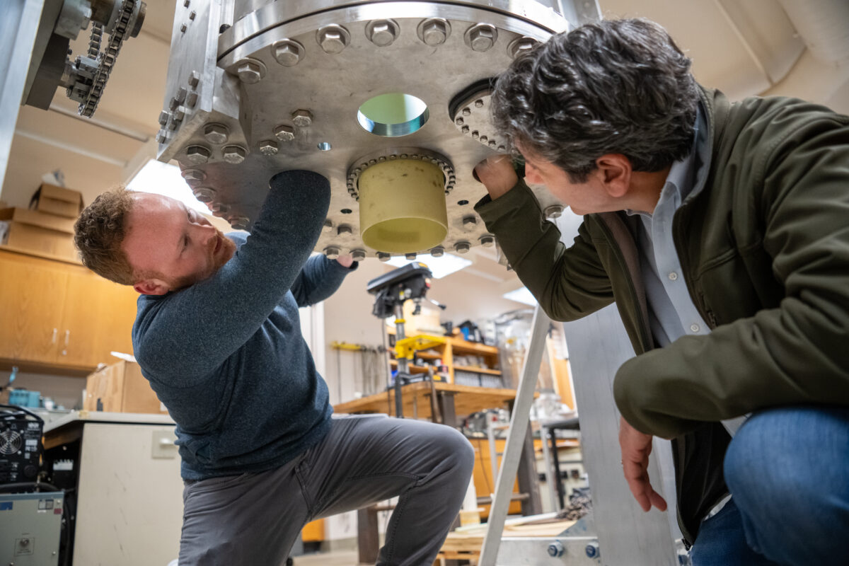 Two men look work under a large metal cylinder housing the magnet.