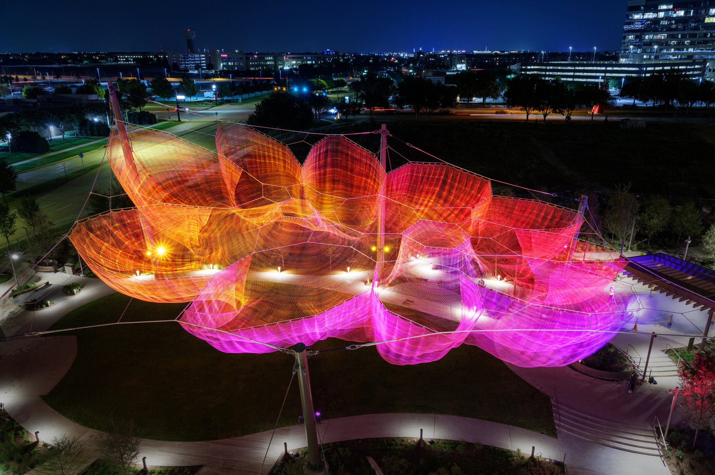 An aerial night view of a large, illuminated orange and pink net sculpture suspended over a walkway.