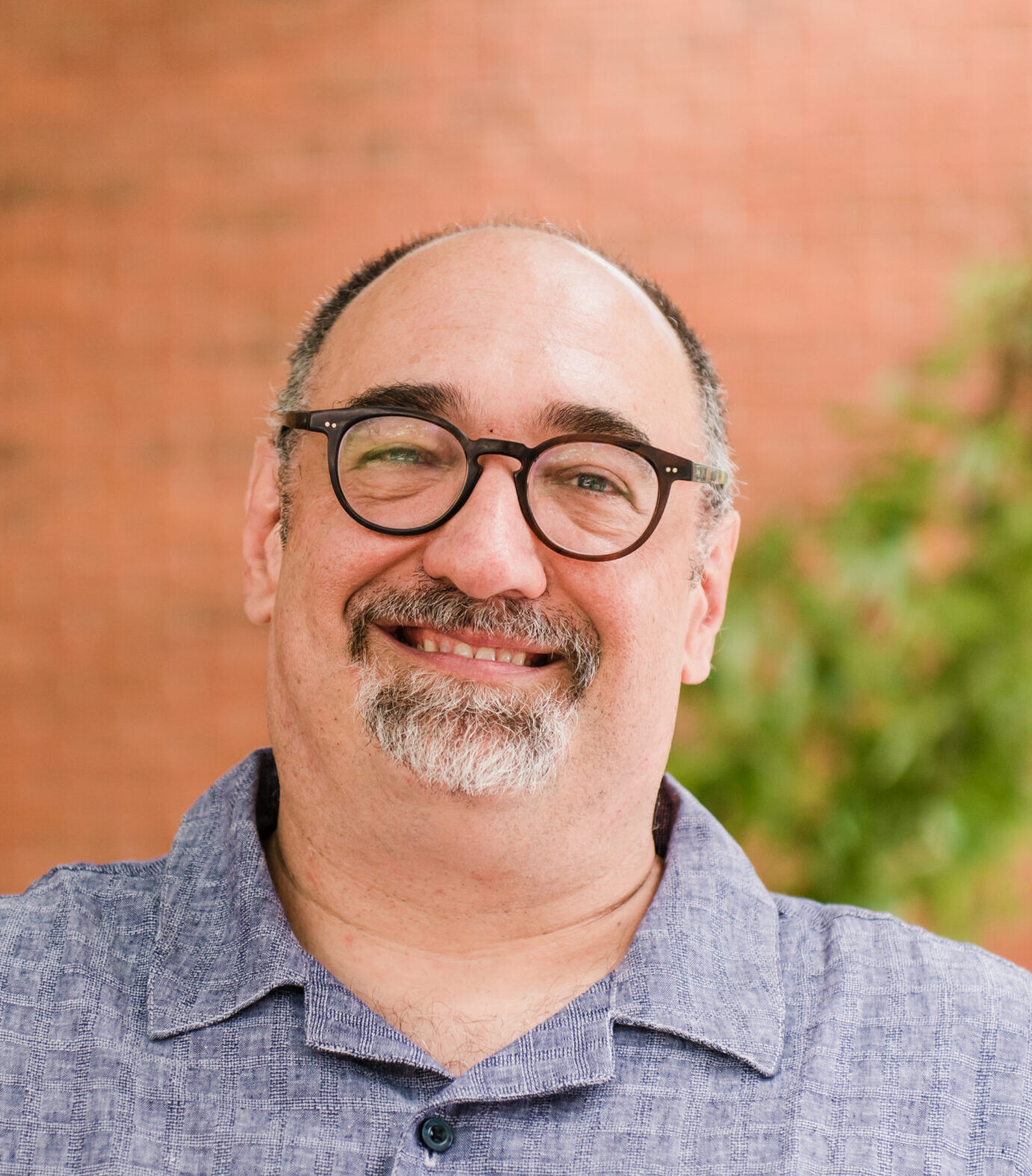 A headshot of a man with a greying beard and dark rimmed glasses wearing a grey collar shirt standing in front of a brick wall and some shrubs