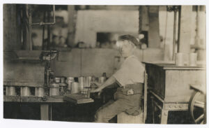 A person sits at a machine operating a tool to process metal cans in a factory setting.