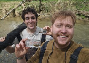 two people holding clear plastic containers each holding a fish, backed by a stream scene