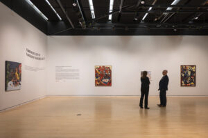 Two people stand in an art gallery looking at artworks hung on white walls.