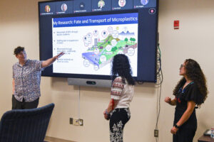 three people present at the front of a conference room; slides on a screen, one pointing to the screen