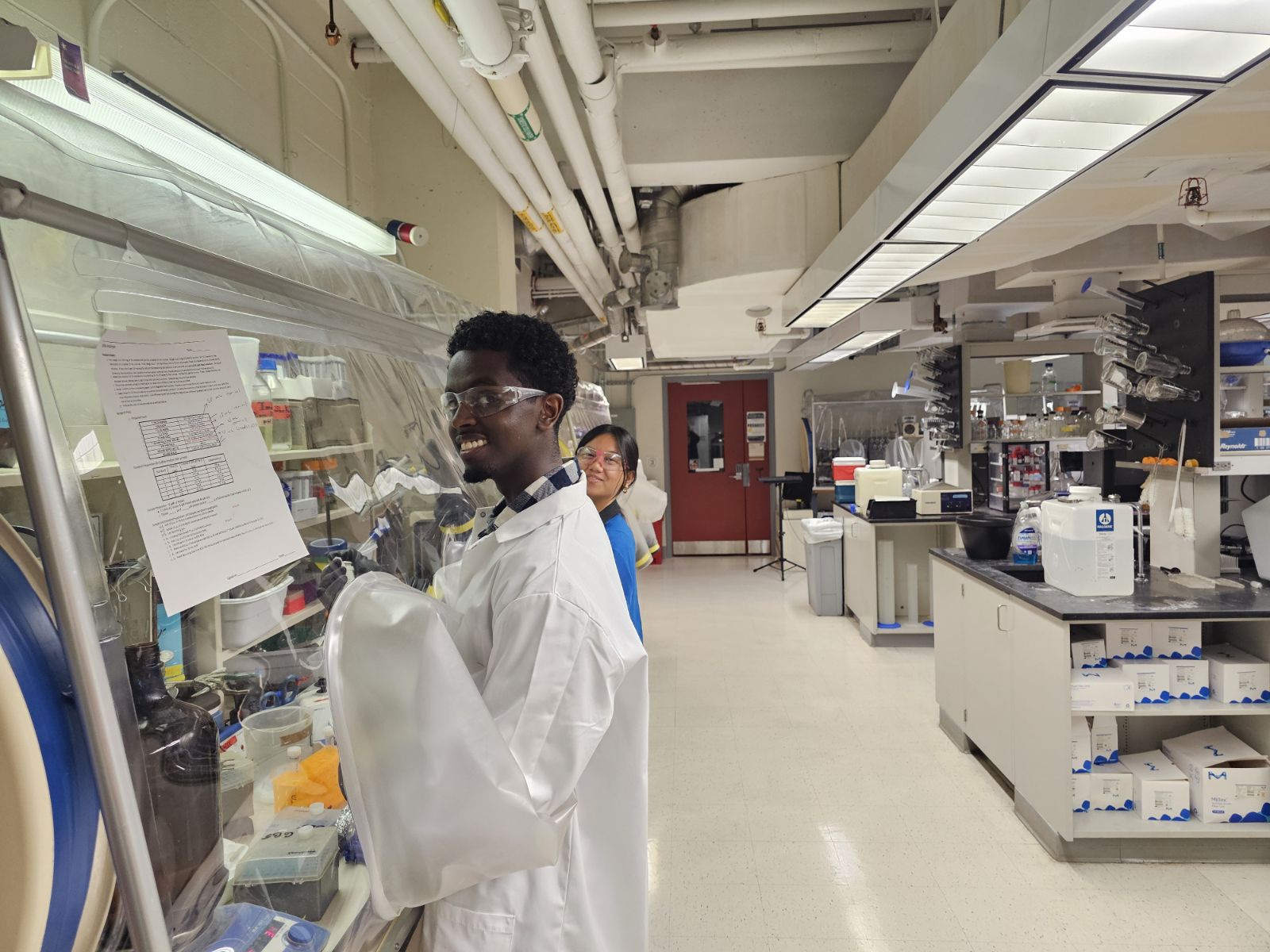Nahom Getachew, and HHMI scholar, works at a fume hood in a lab, wearing a lab coat, gloves, and safety glasses